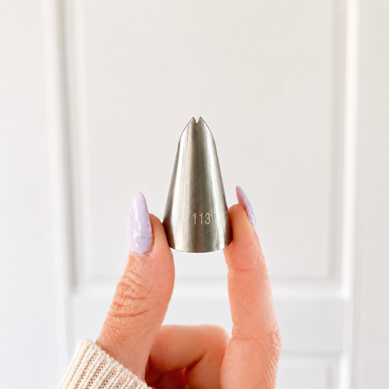 A hand with lavender-painted fingernails holds a stainless steel icing tip with the number 113 engraved on it, set against a soft, out-of-focus white paneled background.