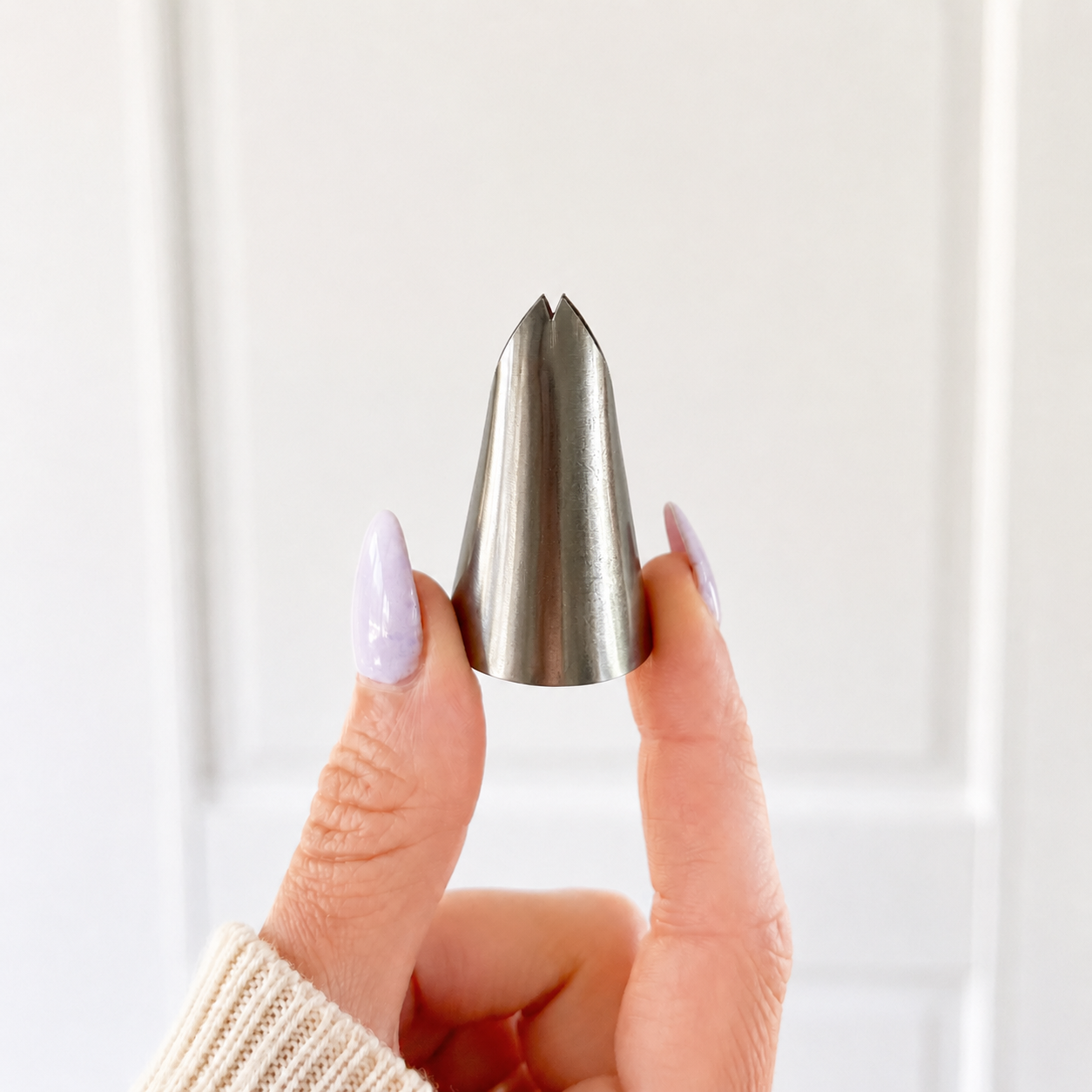 A hand with lavender-painted nails holds a stainless steel piping tip against a soft white paneled background.
