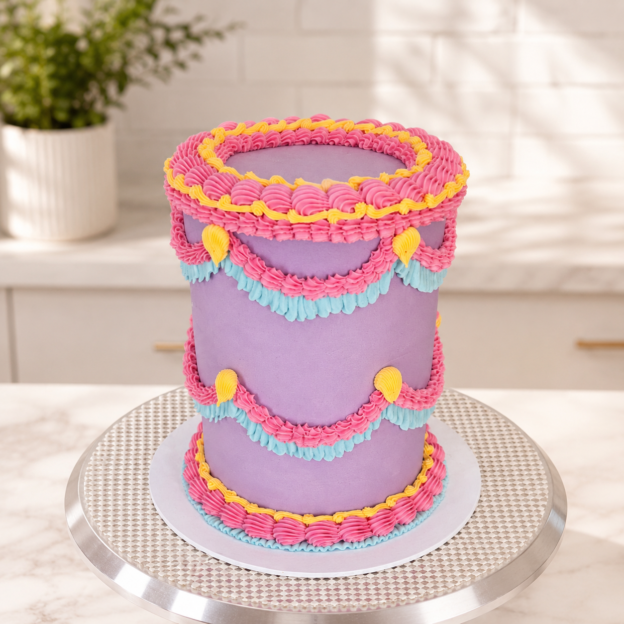 A purple frosted cake decorated with pink, light blue, and yellow piped borders and heart-shaped accents, displayed on a round silver cake stand in a bright kitchen setting.

