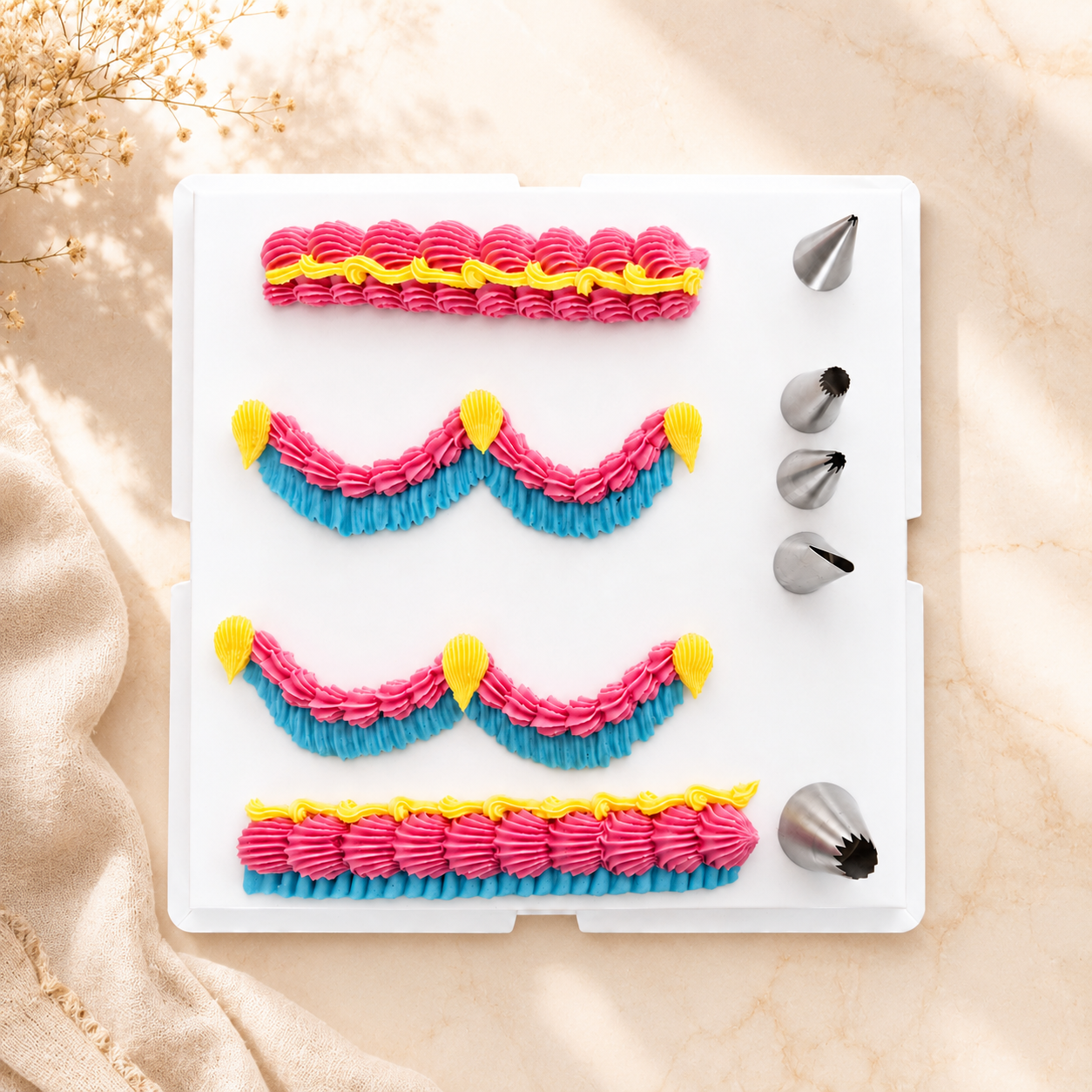 A white plastic piping practice tray with four rows of pink and blue frosting patterns, yellow tip accents, and five metal piping nozzles on the right, photographed on a beige fabric surface with soft sunlight and dried flowers.