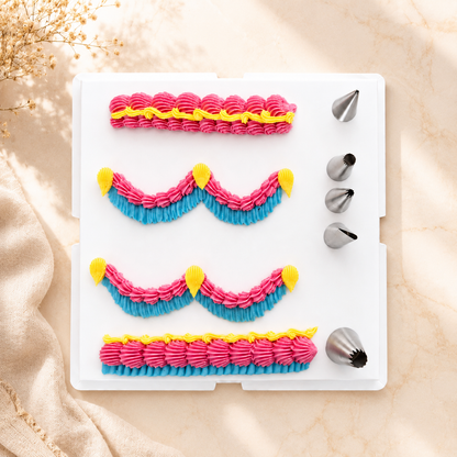 A white plastic piping practice tray with four rows of pink and blue frosting patterns, yellow tip accents, and five metal piping nozzles on the right, photographed on a beige fabric surface with soft sunlight and dried flowers.