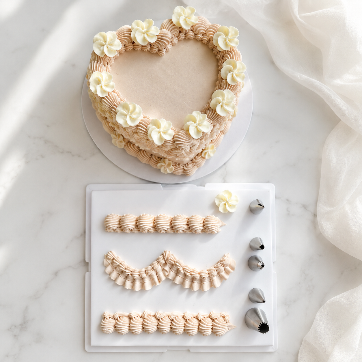 Heart-shaped frosted cake with cream flower decorations on a white round board, shown above a white icing practice sheet with piped patterns and metal piping tips on a marble surface with draped white fabric.