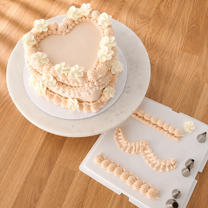 Heart-shaped frosted cake with cream flower decorations on a white round board, shown above a white icing practice sheet with piped patterns and metal piping tips on a marble surface with draped white fabric.