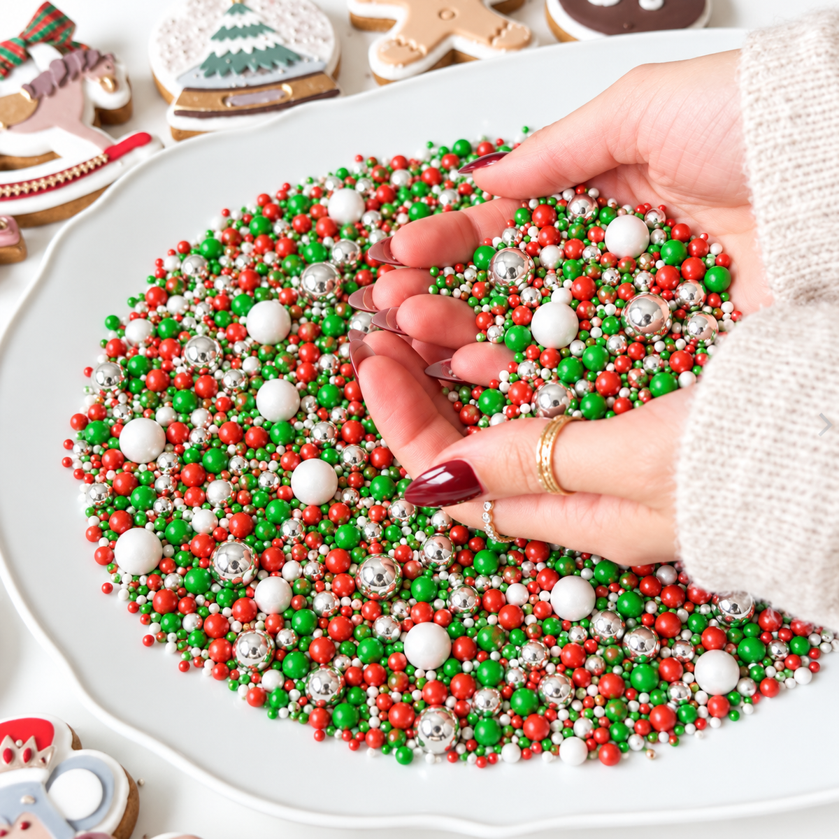 A close-up of hands sprinkling colorful Christmas-themed sprinkles, including red, green, white, and silver ball sprinkles, onto a white plate. The background shows decorated gingerbread cookies.