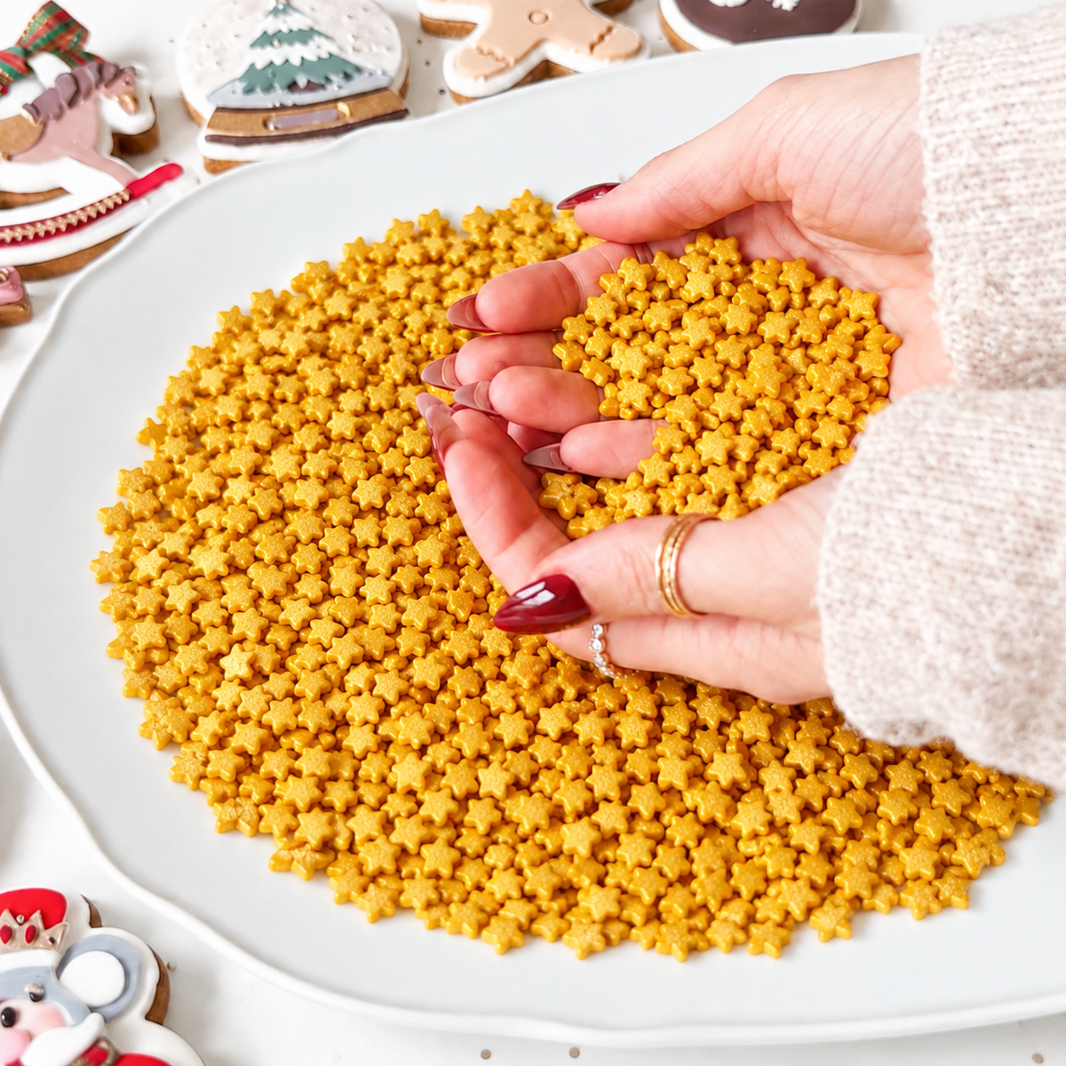 A close-up of a person's hands holding a small pile of golden-yellow star-shaped sprinkles over a large white plate filled with more of the same sprinkles. The background shows some decorated holiday cookies on a white surface.