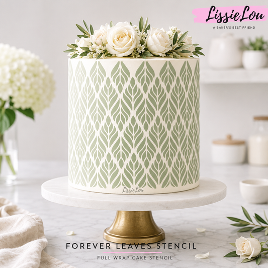 A decorated cake on a white marble and gold pedestal stand, featuring a green and white leaf pattern and topped with white flowers. The background is a softly blurred kitchen setting with white flowers and jars.