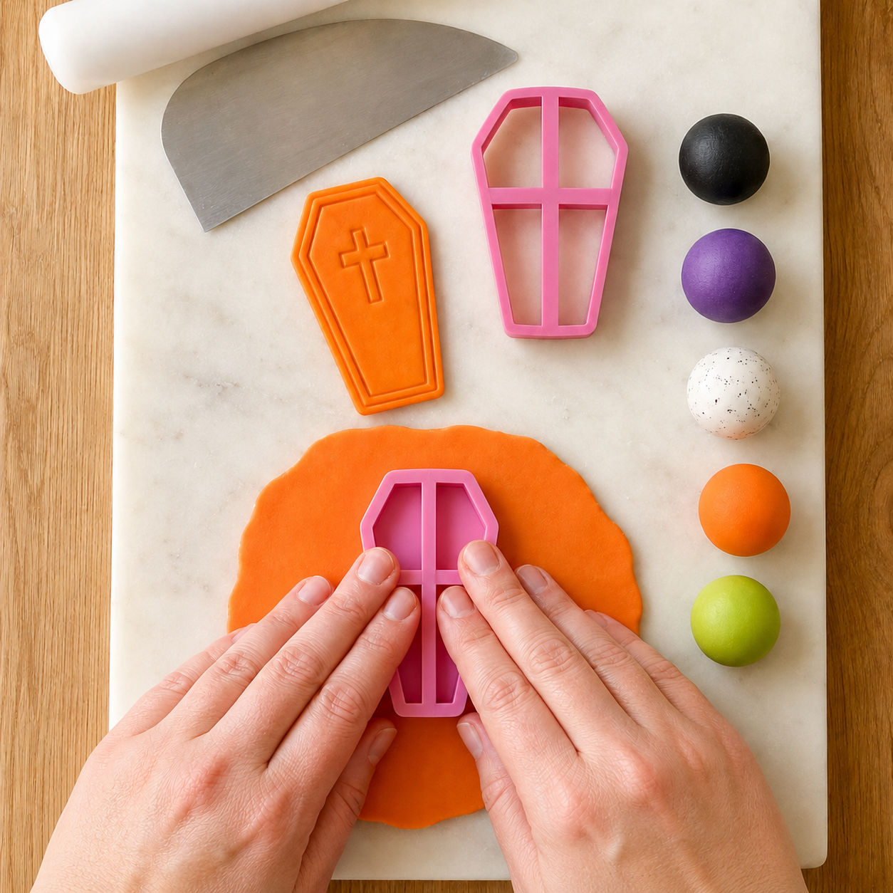 Hands using a pink coffin-shaped cookie cutter on orange dough with Halloween-themed tools and colors.
