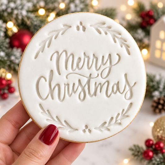 A round white frosted cookie with 'Merry Christmas' written in elegant cursive and decorated with a leafy wreath border, held by a hand with red nail polish against a festive Christmas background with lights and ornaments.