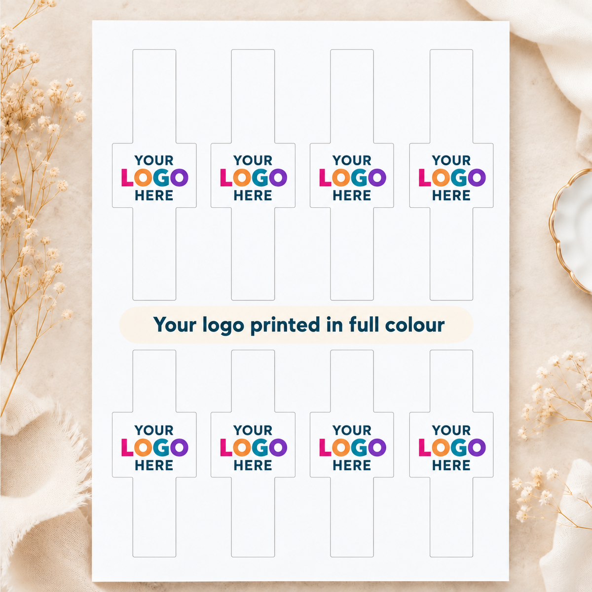 A white sheet showing ten placeholder logo print areas with the text 'YOUR LOGO HERE' and 'Your logo printed in full colour,' styled on a beige surface with dried flowers and soft fabric props.