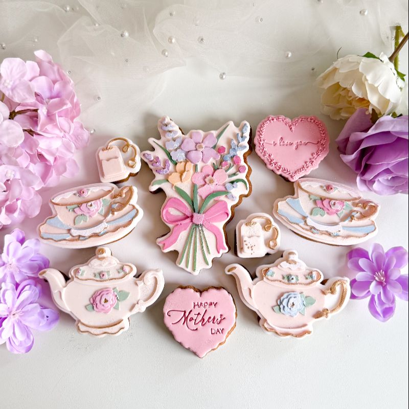 Decorative cookies shaped like teapots, flowers, and hearts with 'Happy Mother's Day' text, surrounded by flowers on a light background.