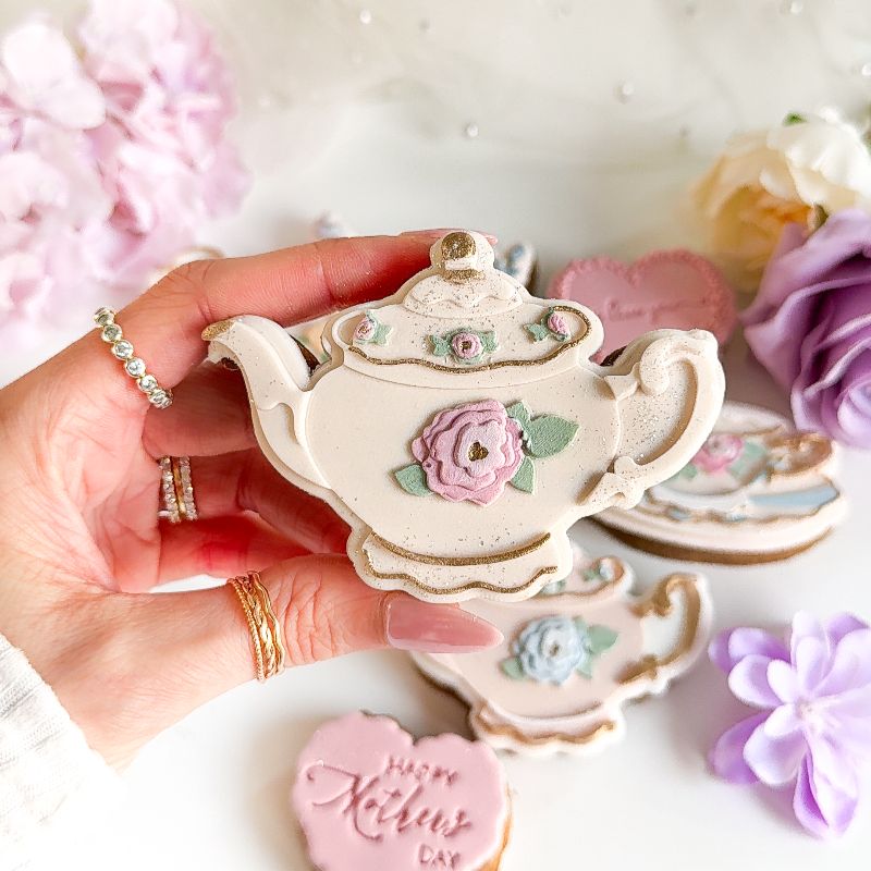 Decorative teapot-shaped cookie held by a hand with floral and cookie decorations in the background.