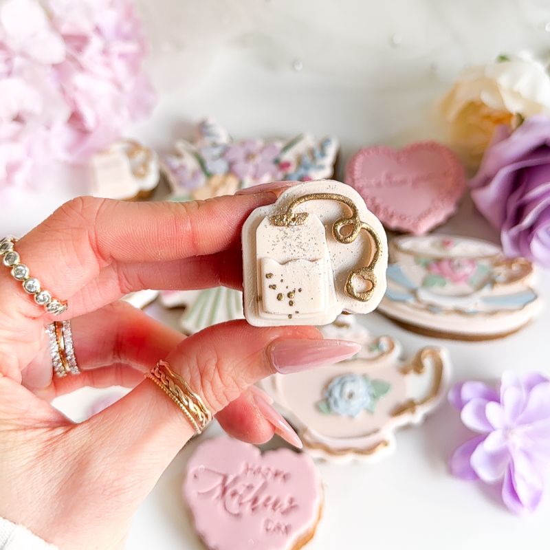 Hand holding a decorative cookie with a teacup design, surrounded by other decorated cookies on a white surface.