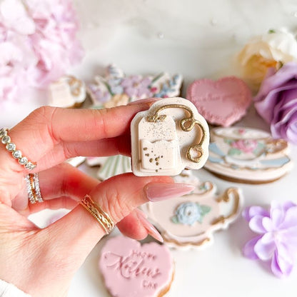 Hand holding a decorative cookie with a teacup design, surrounded by other decorated cookies on a white surface.