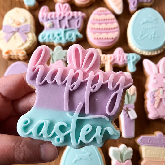 Hand holding a 'Happy Easter' cookie with more decorated cookies in the background.