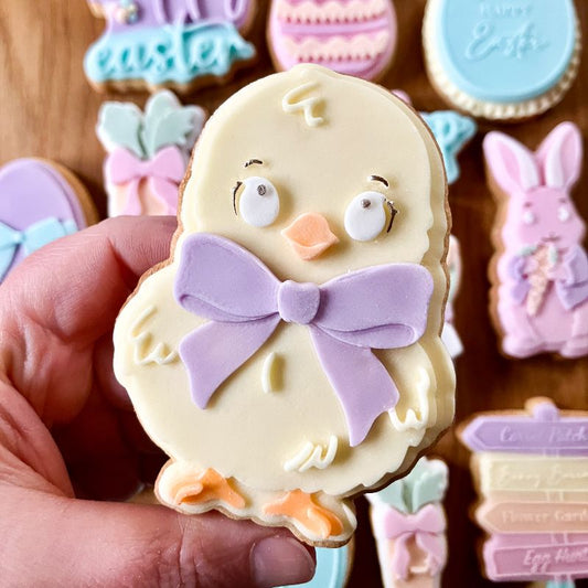 Hand holding a decorated cookie shaped like a chick with a purple bow, surrounded by other Easter-themed cookies.
