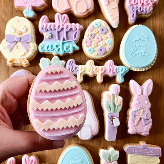Decorative Easter cookies on a wooden surface with a hand holding a pink egg-shaped cookie.