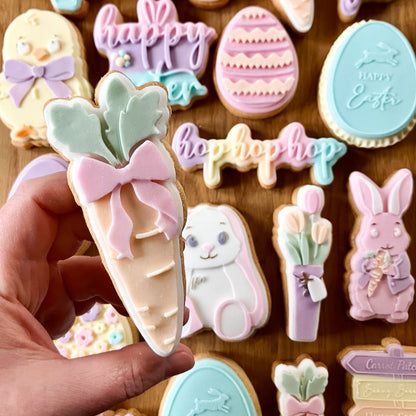 Hand holding a pastel-colored carrot-shaped cookie with Easter-themed cookies in the background.